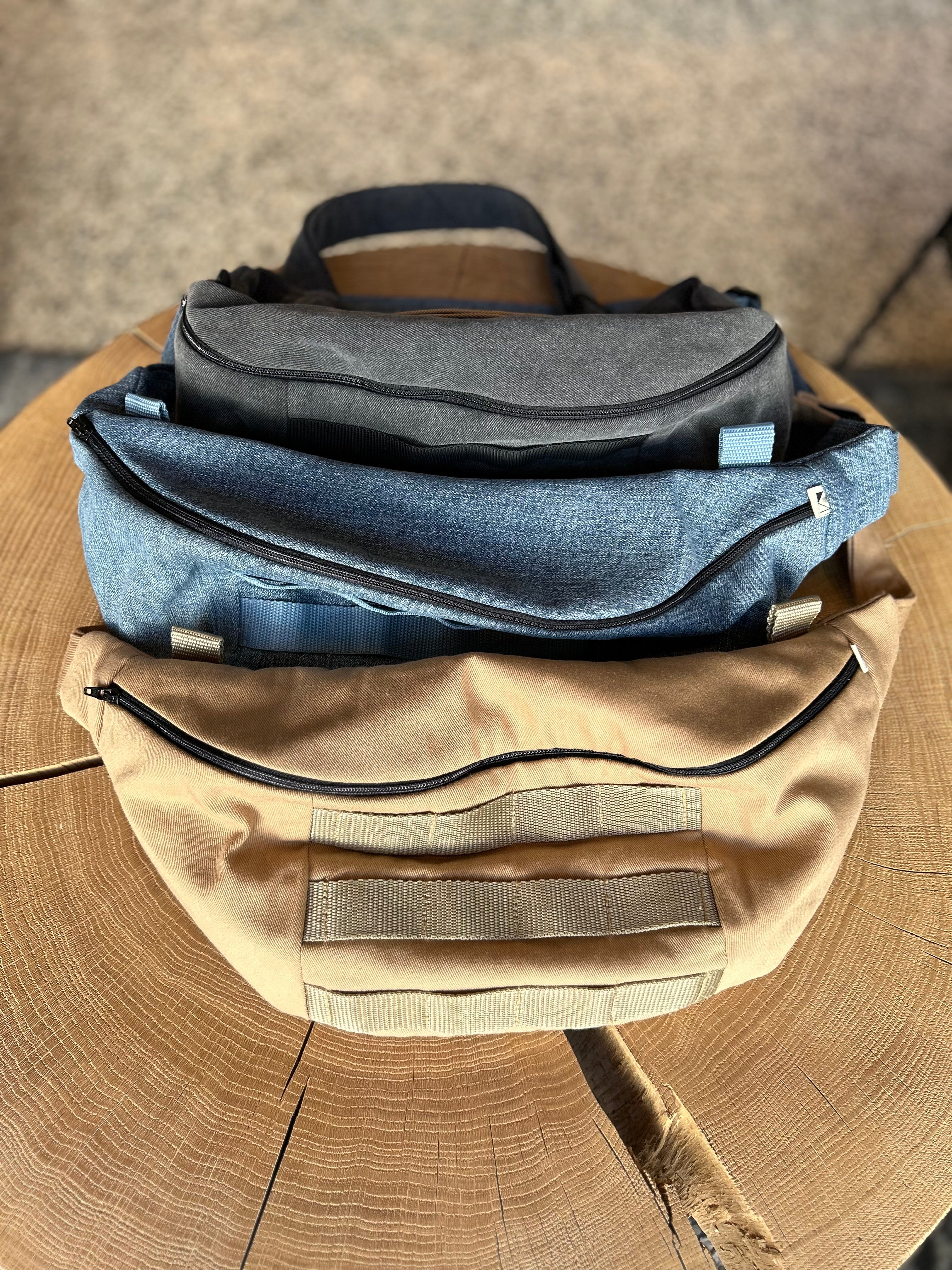 3 Taschen in Form einer Banane in der Farbe Blau, Beige und Grau, liegen auf einem Holztisch