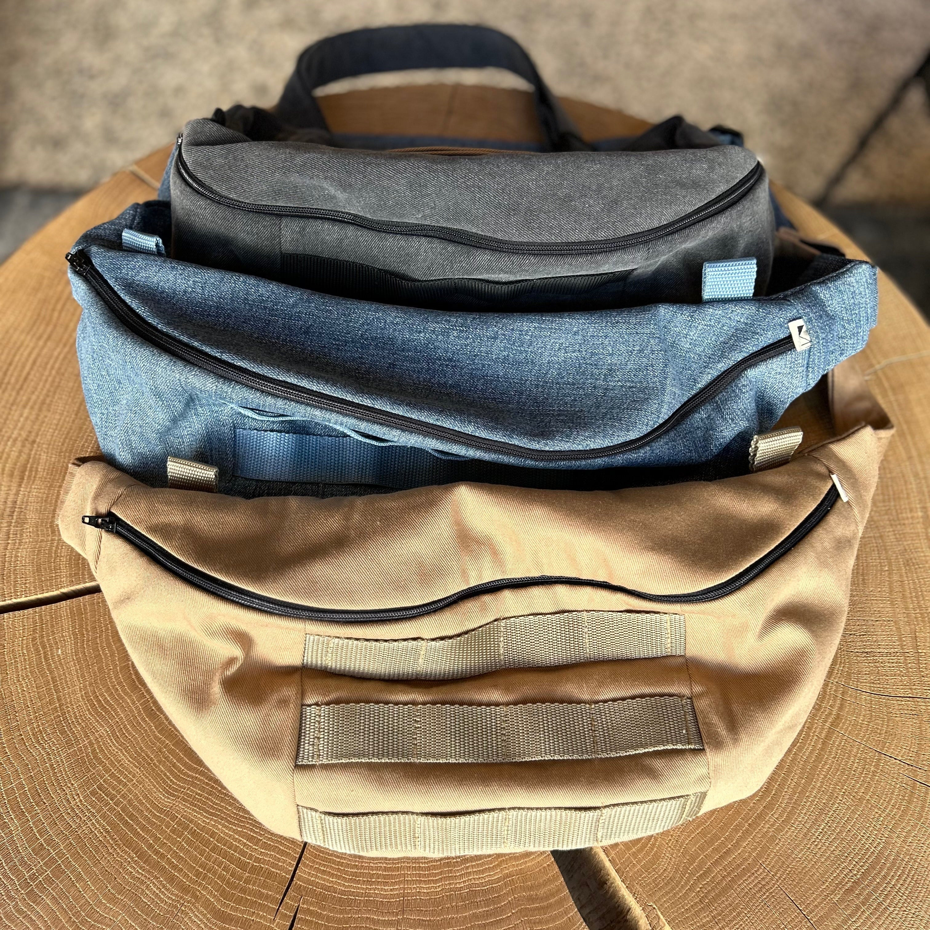 3 Taschen in Form einer Banane in der Farbe Blau, Beige und Grau, liegen auf einem Holztisch