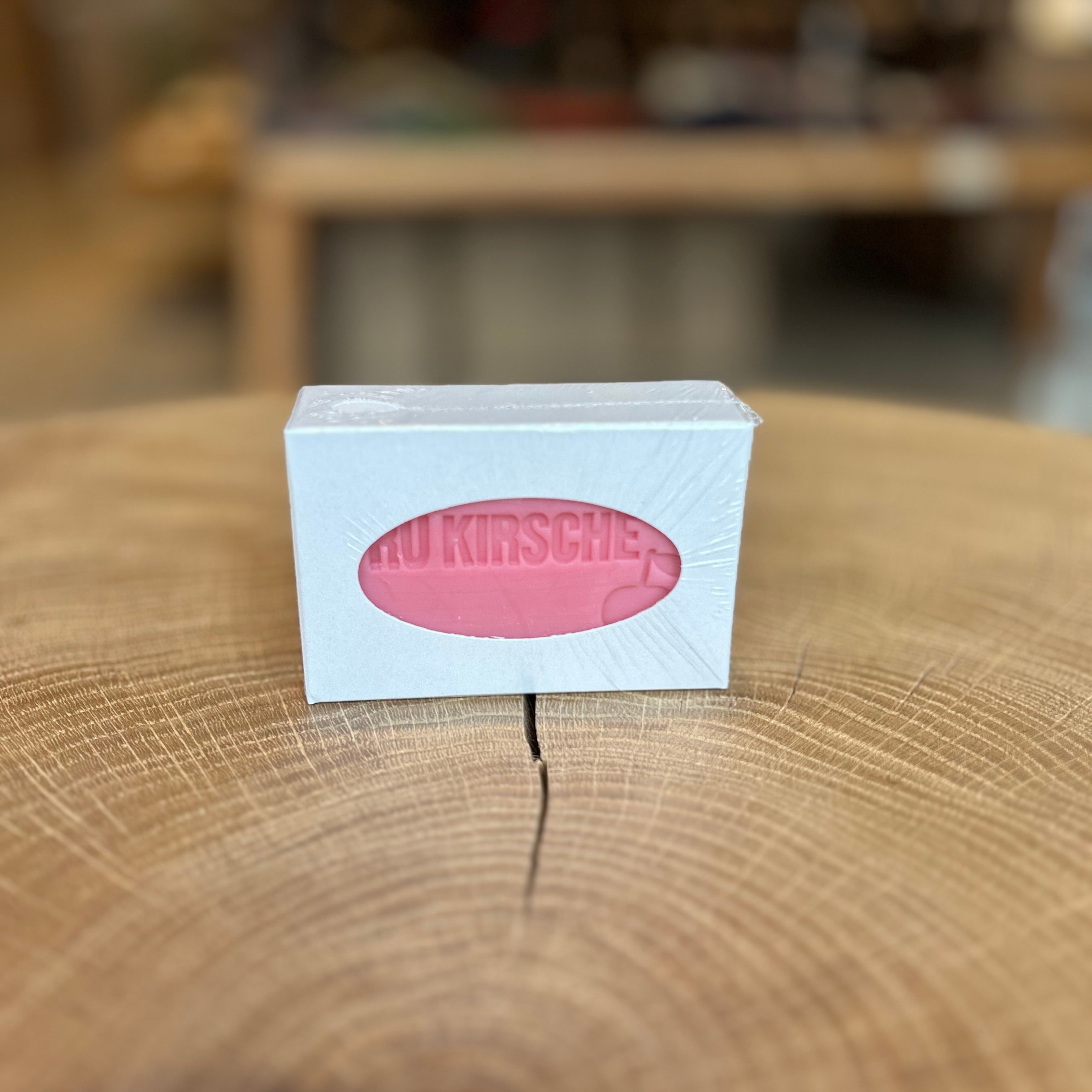 RÜ Kirsche Blockseife in weißer Verpackung mit Sichtfenster, rosa Seifenstück sichtbar. Präsentiert auf dekorativem Holztisch, im Hintergrund unscharfe Boutique-Atmosphäre. Handgemachte Kirschduft-Seife aus Essen-Rüttenscheid – ideal als Geschenk, Duftseife oder pflegende Handseife.