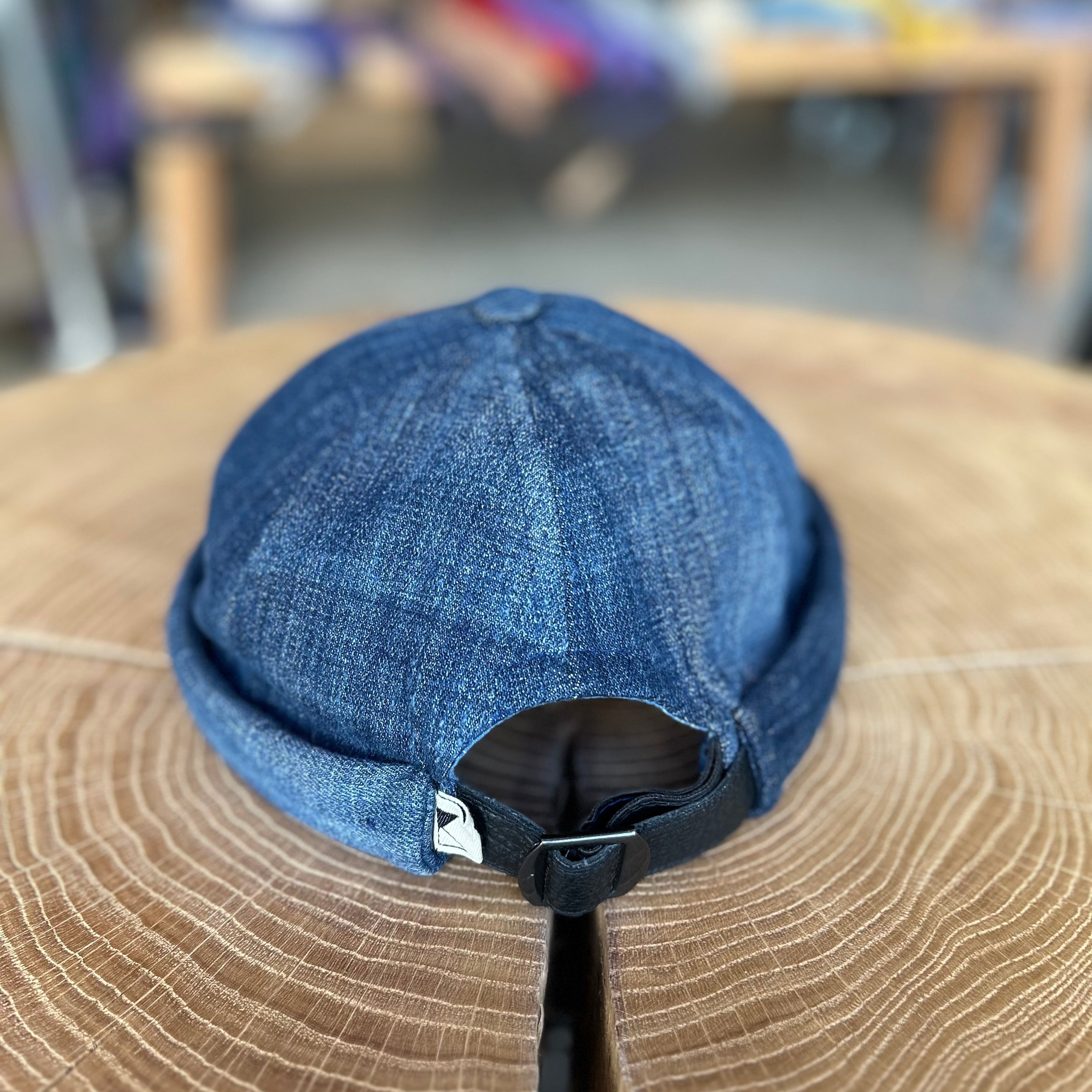 Fischermütze in der Farbe Dunkel Jeans, Rückseite der Fischermütze, liegt auf einem Holztisch