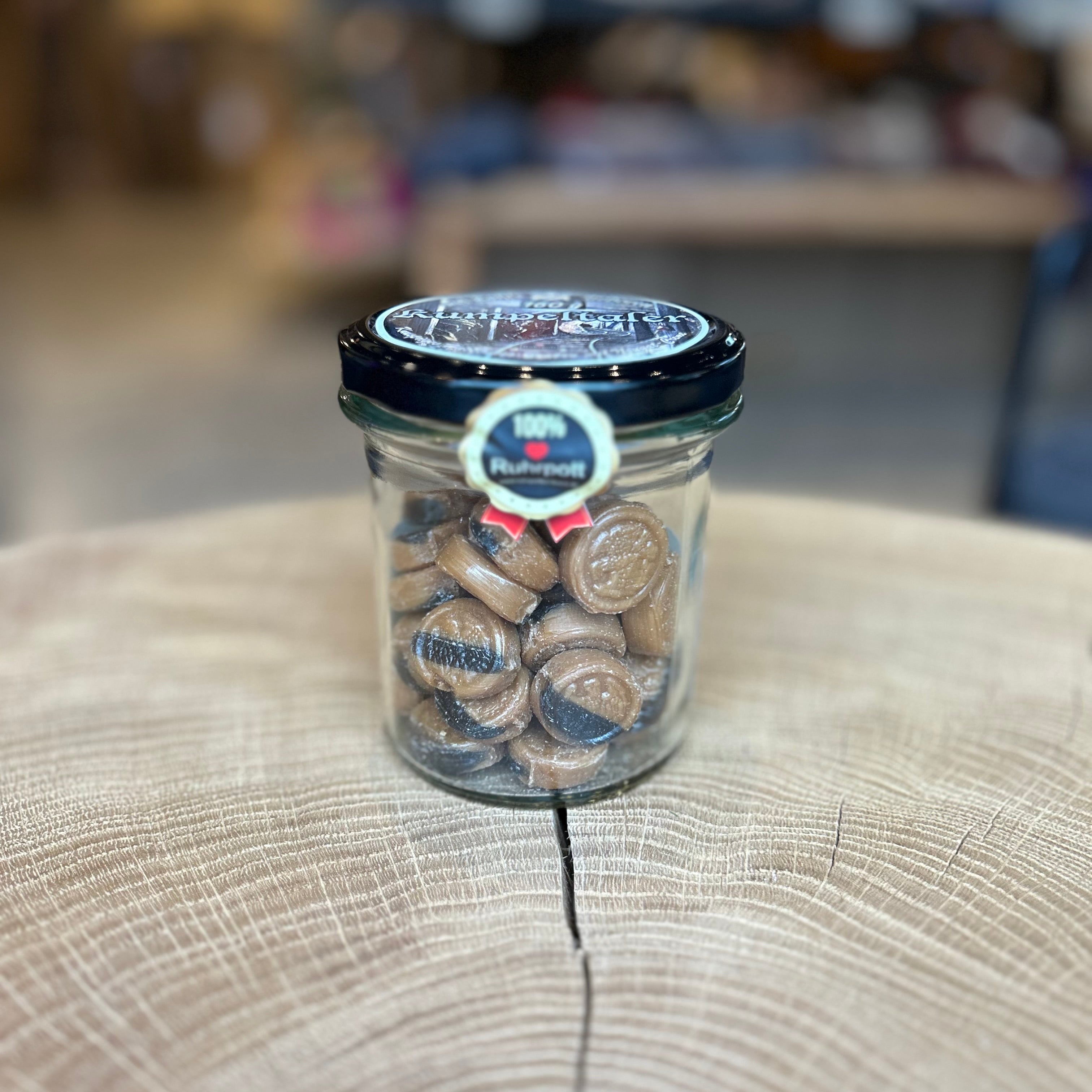 Glas mit braunen „Kumpeltaler Nougat Bonbons“ auf einem Holzuntergrund. Das Schraubglas ist gefüllt mit runden Nougatbonbons und trägt ein Etikett mit der Aufschrift „100% Ruhrpott“. Ideal als Geschenk oder regionales Mitbringsel aus dem Ruhrgebiet.