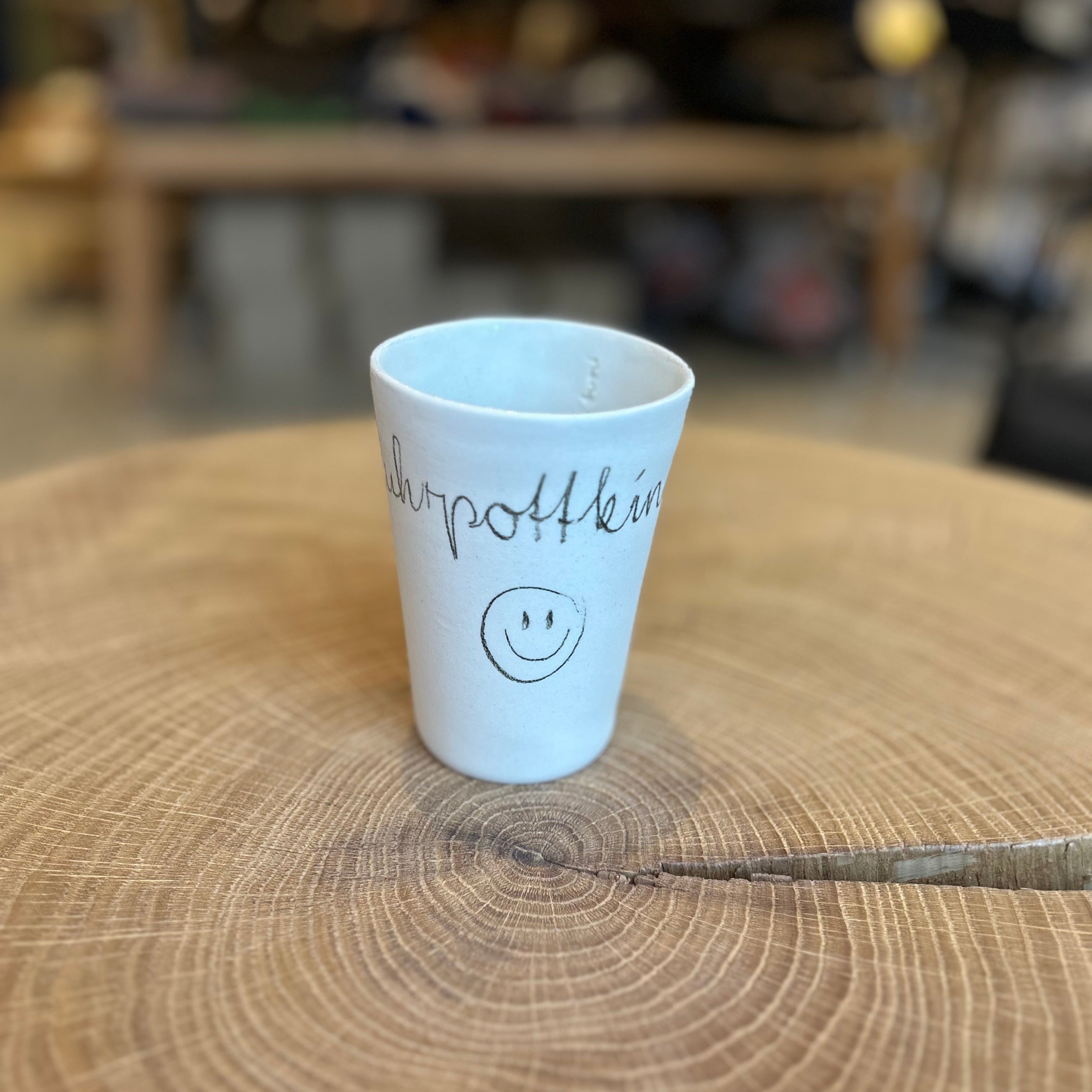 Handgefertigte Keramikbecher in unterschiedlichen Größen mit der Aufschrift „Ruhrpottkind“ und Smiley, auf einer rustikalen Holzscheibe arrangiert – minimalistisches Design, regionale Keramik, Geschenkidee aus dem Ruhrgebiet.