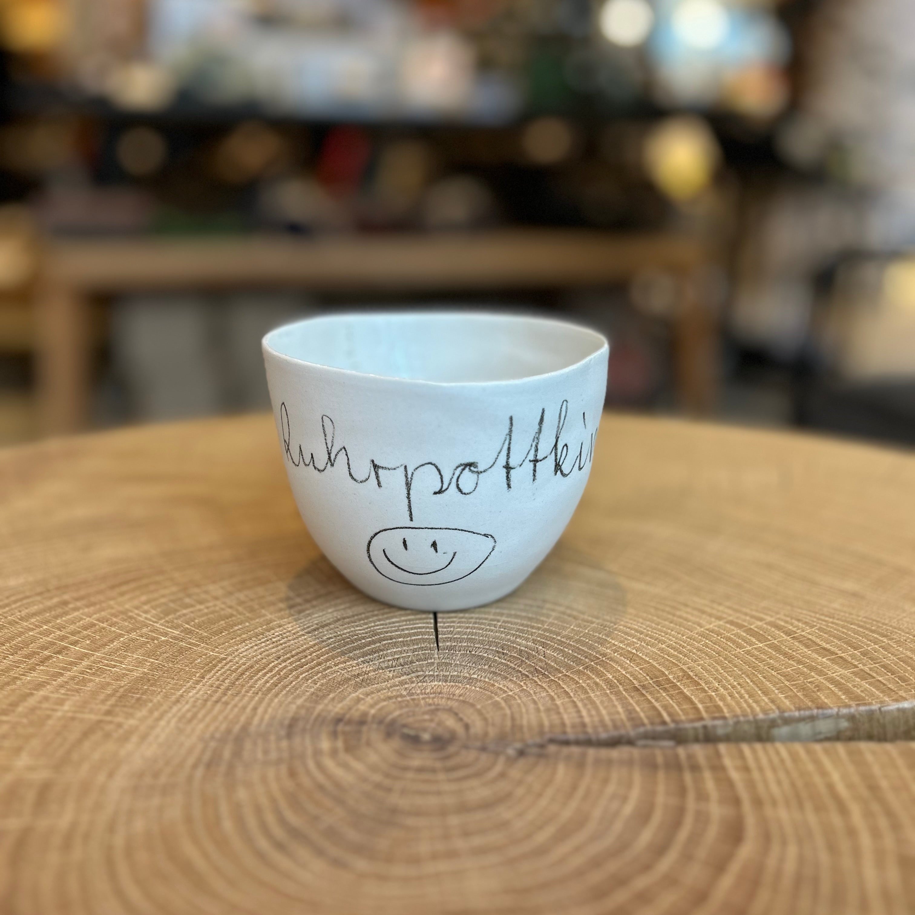 Handgefertigte Keramikschale „Ruhrpottkind“ von Manufaktur Peng, kleine weiße Bowl mit handgeschriebener Schrift und Smiley, minimalistisches Design, Unikat aus Keramik auf Holzoberfläche.