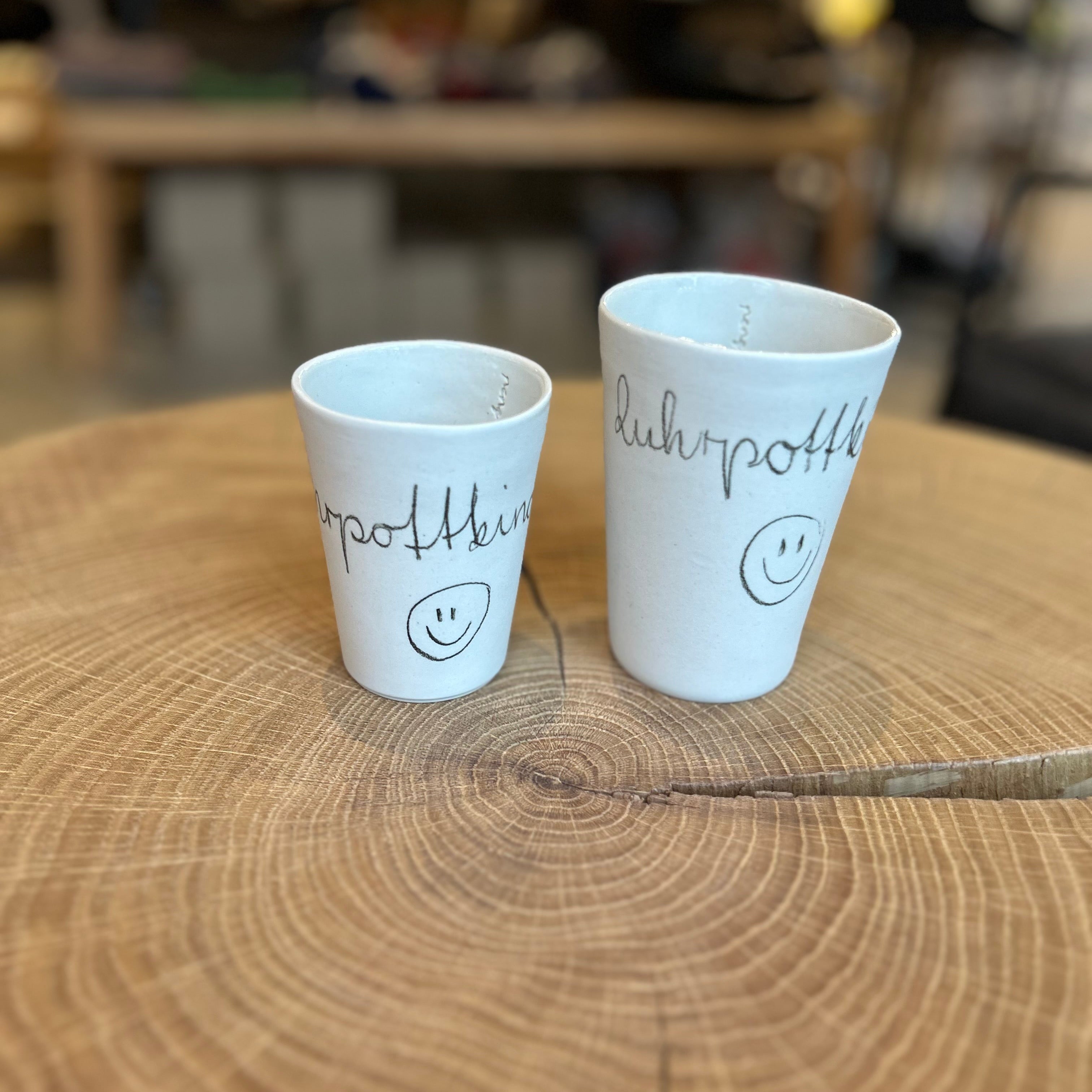Zwei handgefertigte Keramikbecher in unterschiedlichen Größen mit der Aufschrift „Ruhrpottkind“ und Smiley, auf einer rustikalen Holzscheibe arrangiert – minimalistisches Design, regionale Keramik, Geschenkidee aus dem Ruhrgebiet.