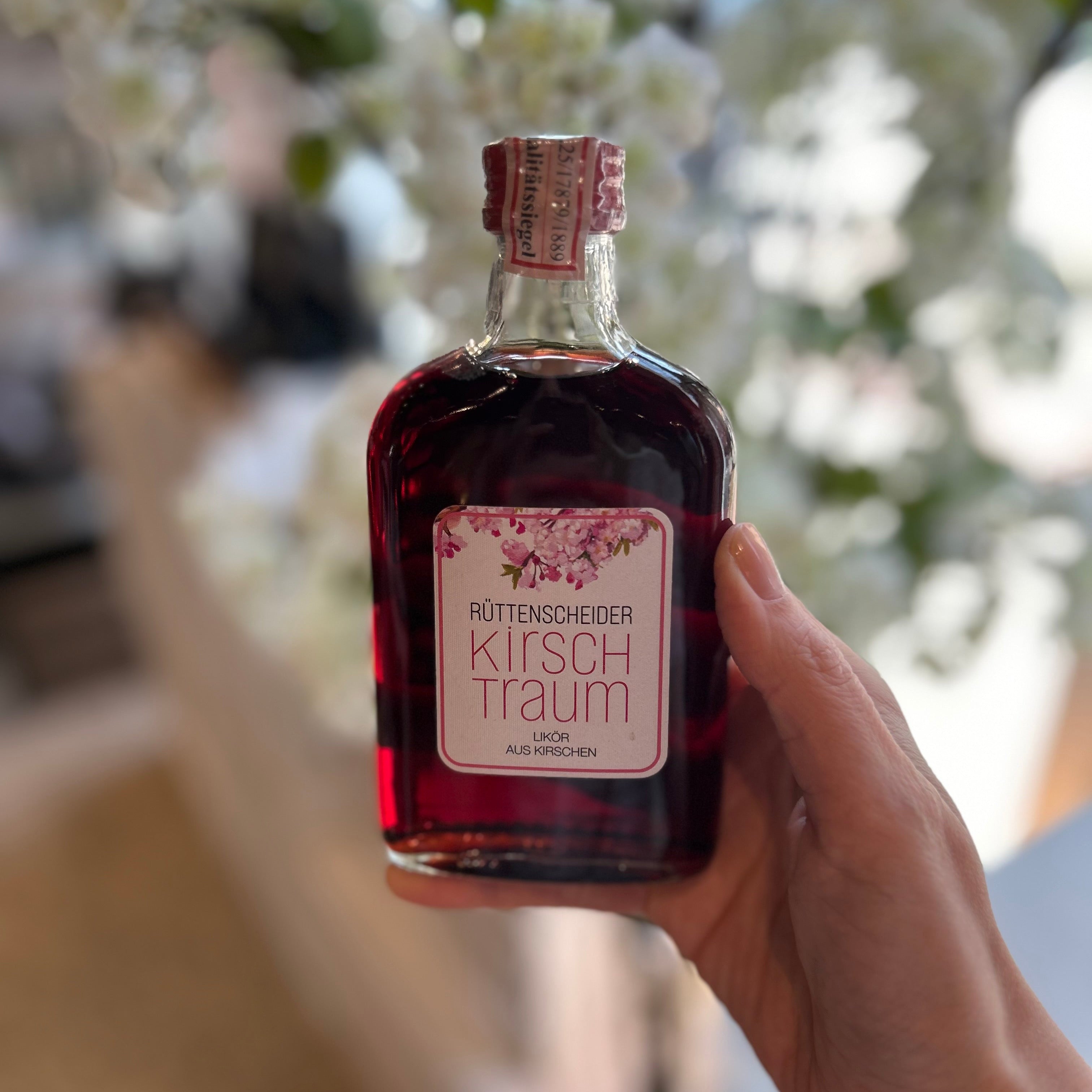 Rüttenscheider „Kirschtraum“ Kirschlikör in Glasflasche in klein in der Hand gehalten vor unscharfem Hintergrund – handgemachter Kirschlikör aus Kirschen als Spirituosen-Spezialität.