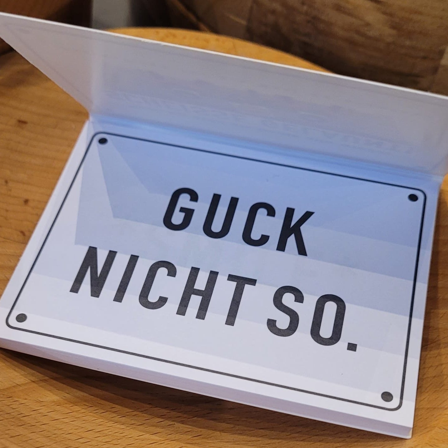 Haftnotizen mit humorvollem Spruch „Boah ey, sind die heute alle wieder scheisse gelaunt!“ auf einem Holztisch. Das Schild zeigt eine Illustration mit drei Figuren, darunter ein Smiley-Gesicht in der Mitte. Lustiges Deko-Schild als Geschenk, Bürodeko oder Tischaufsteller für gute Laune.