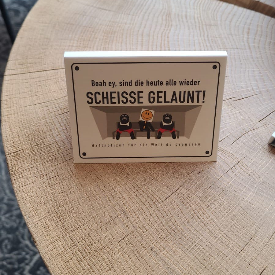 Haftnotizen mit humorvollem Spruch „Boah ey, sind die heute alle wieder scheisse gelaunt!“ auf einem Holztisch. Das Schild zeigt eine Illustration mit drei Figuren, darunter ein Smiley-Gesicht in der Mitte. Lustiges Deko-Schild als Geschenk, Bürodeko oder Tischaufsteller für gute Laune.
