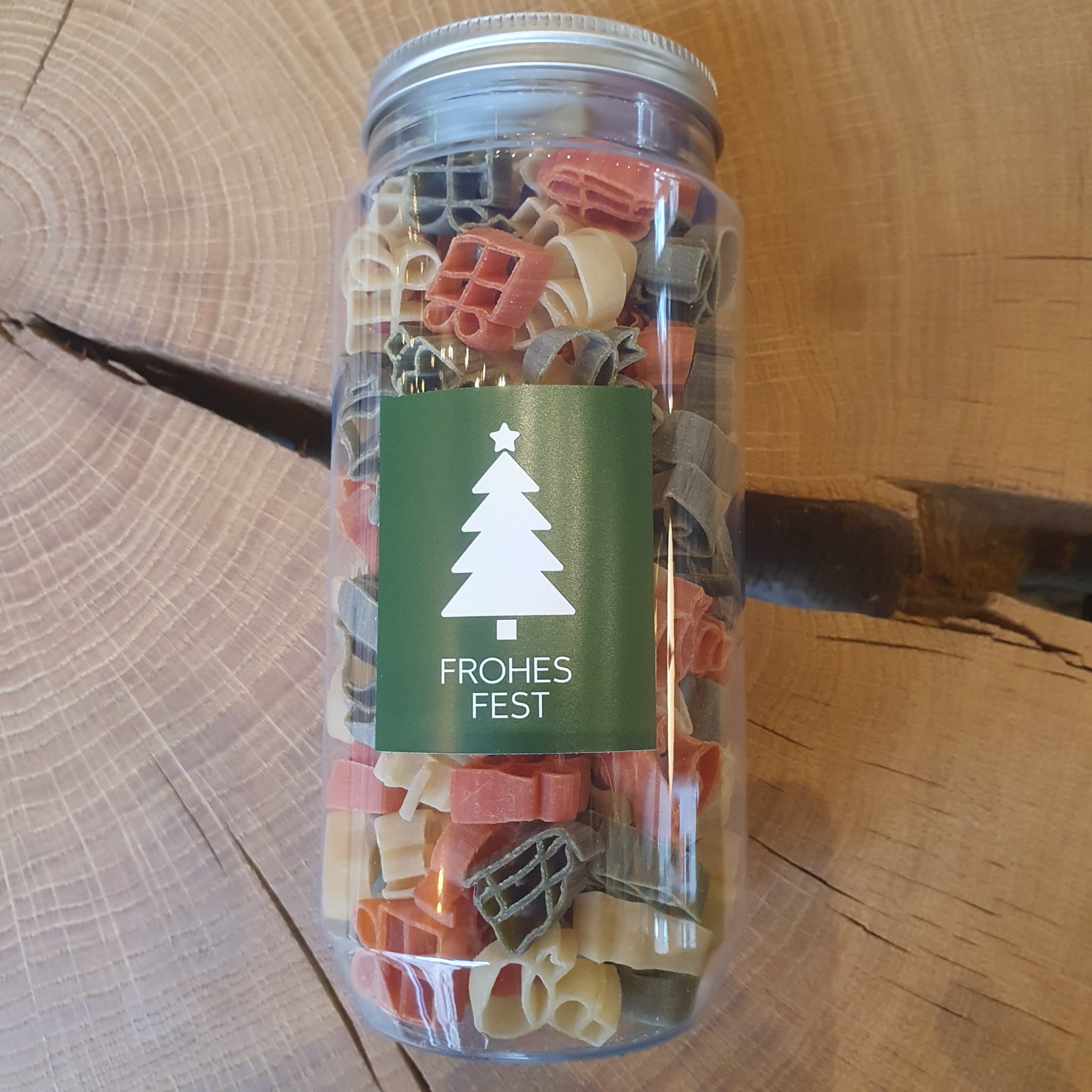 Weihnachtliche bunte Nudeln in Geschenkflasche mit grünem Etikett „Frohes Fest“ und Tannenbaum-Motiv, stehend auf einem rustikalen Holztisch. Weihnachtsnudeln in verschiedenen Formen – perfektes Geschenk, Mitbringsel oder Dekoidee für die Weihnachtszeit.