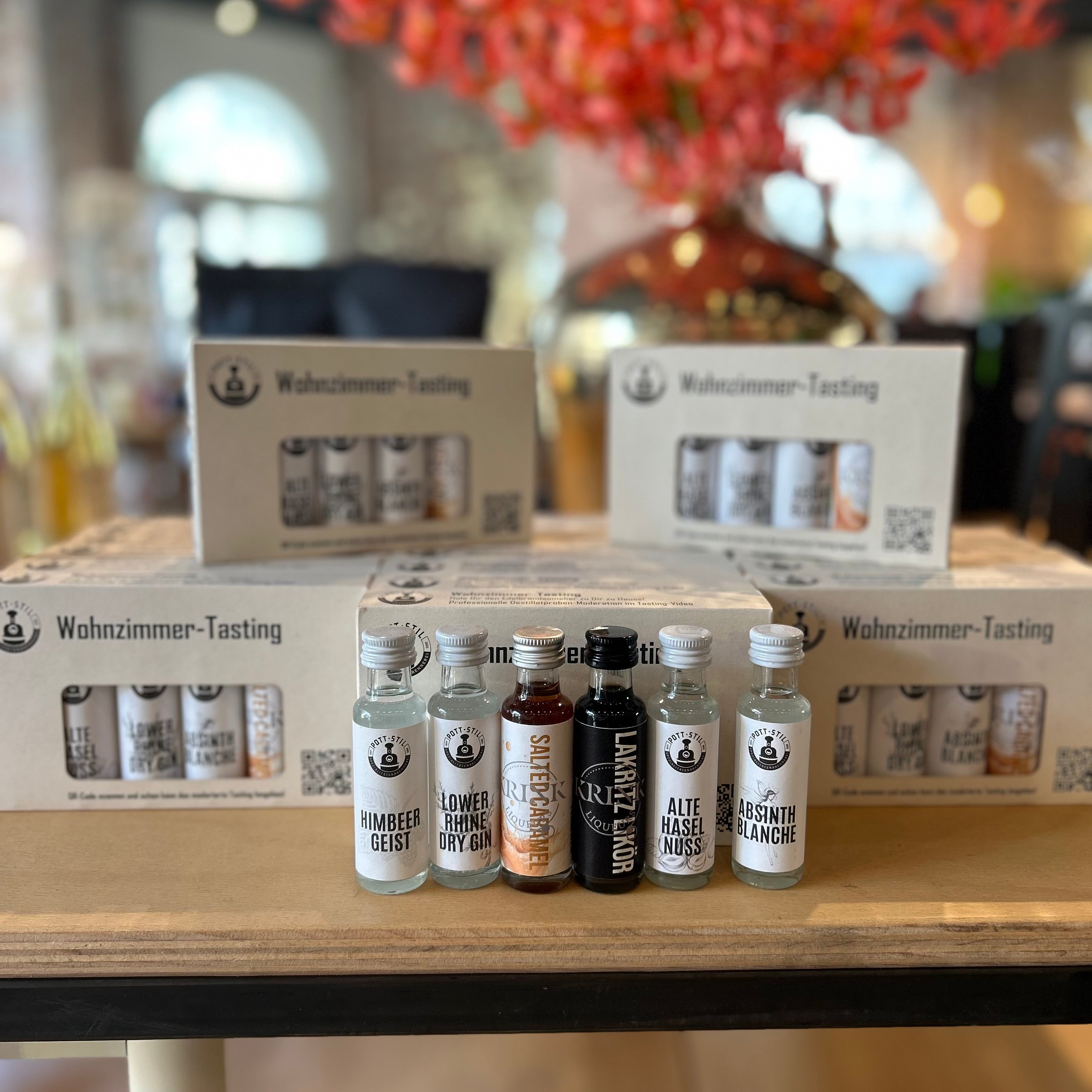 Wohnzimmer-Tasting Set mit verschiedenen Spirituosen-Miniaturen wie Himbeergeist, Gin, Lakritzlikör und Absinth auf einem Holzbrett vor Verpackungen des Wohnzimmer-Tasting Probierpakets – Spirituosen Verkostungsset für Zuhause.