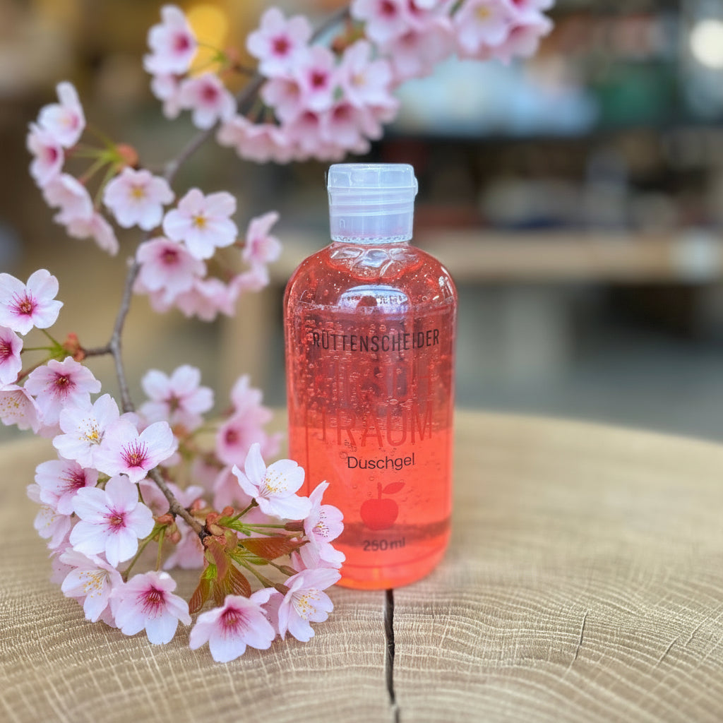 Rüttenscheider Kirschtraum Duschgel in transparenter 250-ml-Flasche mit rotem, kirschfarbenem Gel. Stehend auf einem Holztisch, im Hintergrund unscharfe Boutique- oder Ladenatmosphäre. Pflegendes Duschgel mit fruchtigem Kirschduft – ideal als Geschenk, Wellnessprodukt oder Pflegeartikel aus Essen-Rüttenscheid.