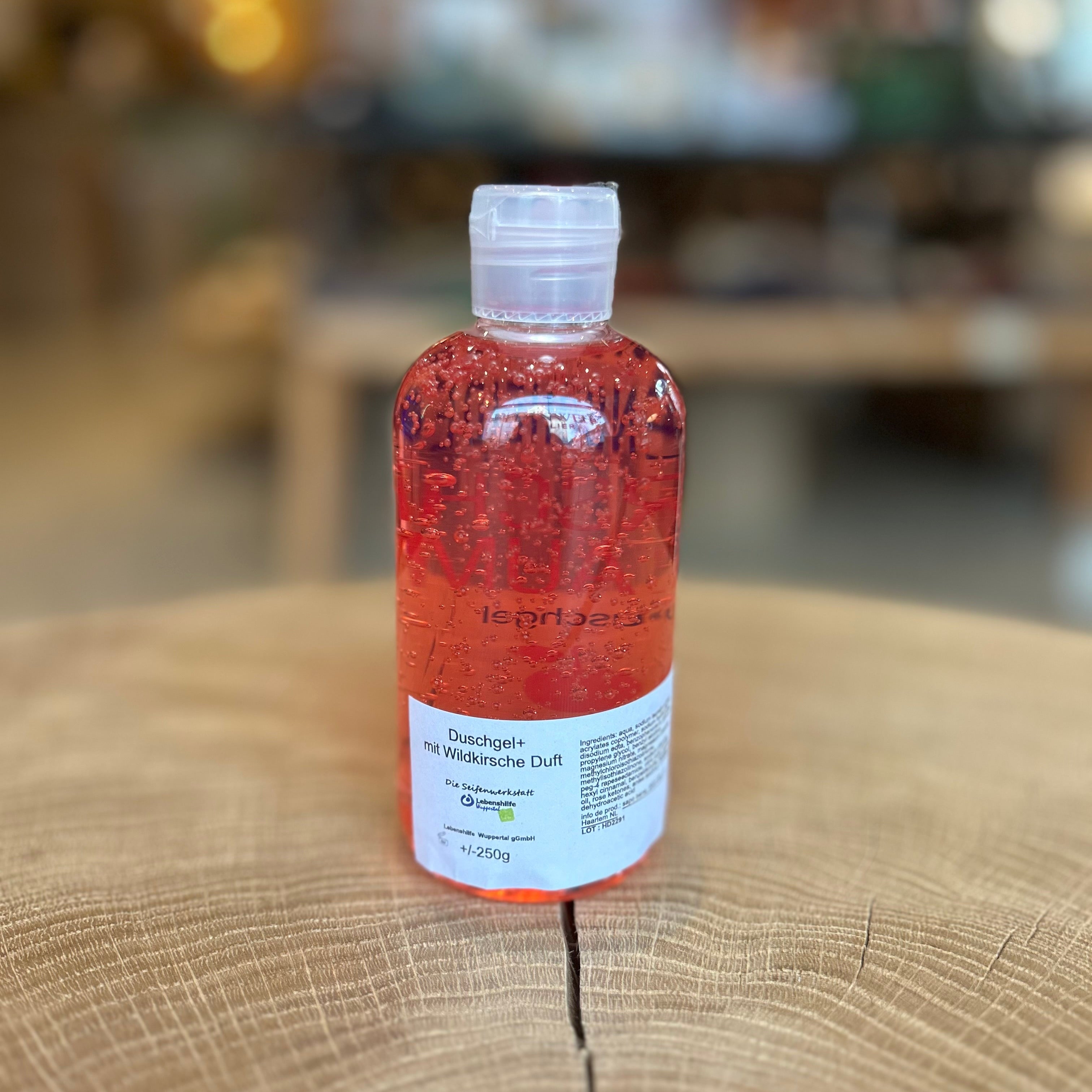 Rüttenscheider Kirschtraum Duschgel in transparenter 250-ml-Flasche mit rotem, kirschfarbenem Gel. Stehend auf einem Holztisch, im Hintergrund unscharfe Boutique- oder Ladenatmosphäre. Pflegendes Duschgel mit fruchtigem Kirschduft – ideal als Geschenk, Wellnessprodukt oder Pflegeartikel aus Essen-Rüttenscheid.