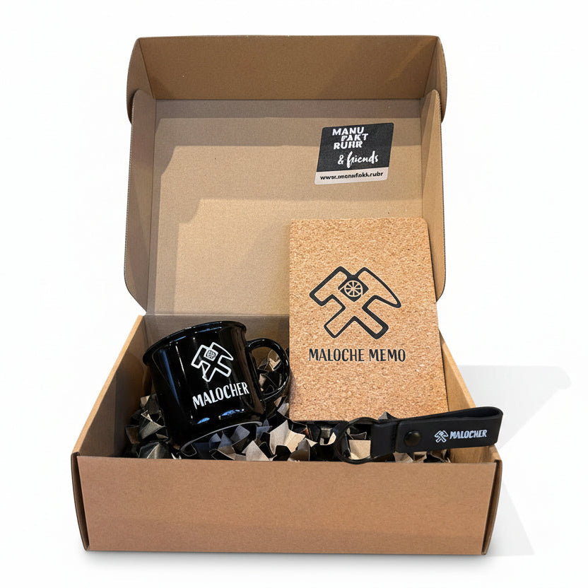 Offene Geschenkbox mit Ruhrpott-Motiv „Malocher“: schwarze Emaille-Tasse, Kork-Notizbuch „Maloche Memo“ und Schlüsselanhänger, stilvoll verpackt in Geschenkbox von Manufakt Ruhr & friends.