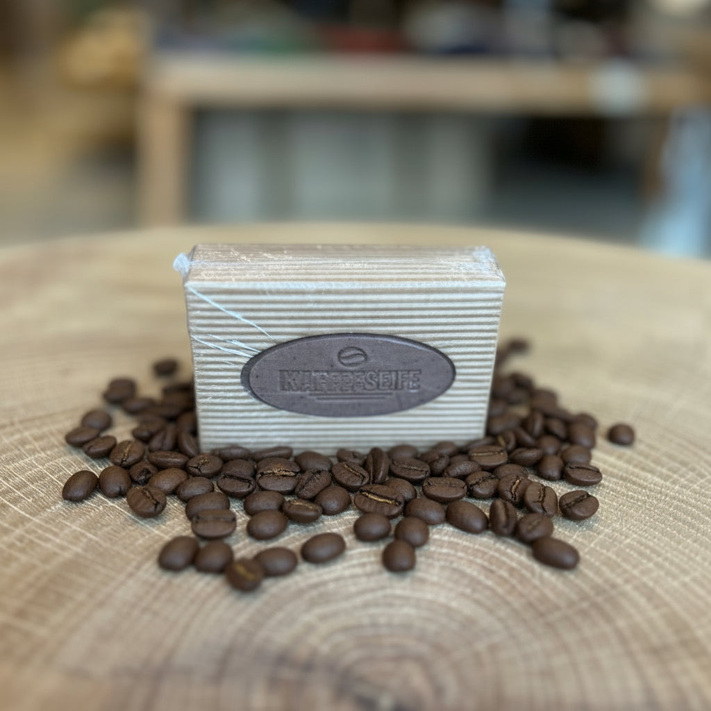 Kaffeeseife im Block, verpackt in naturfarbener Papierbanderole mit ovalem Etikett „Kaffeeseife“. Präsentiert auf einem dekorativen Holztisch, im Hintergrund unscharfe Ladenatmosphäre. Handgemachte Peelingseife mit Kaffee – ideal zur Geruchsneutralisation und als natürliche Küchen- oder Handseife.