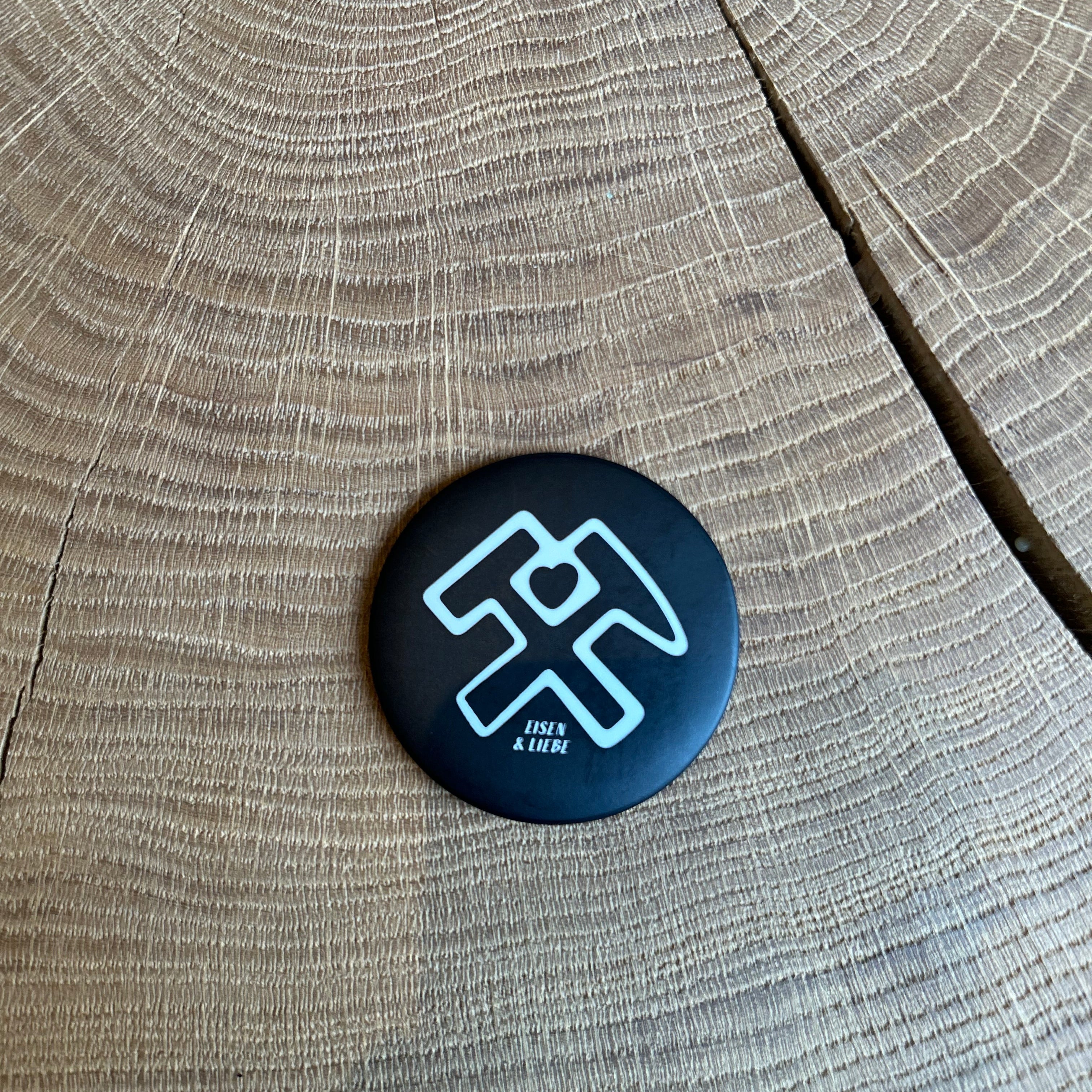 Runder schwarzer Magnet mit weißem Hammer-und-Schlägel-Symbol und Herz sowie der Aufschrift „Eisen & Liebe“ auf einer Holzoberfläche – Bergbau Symbol als Deko- oder Geschenkartikel.