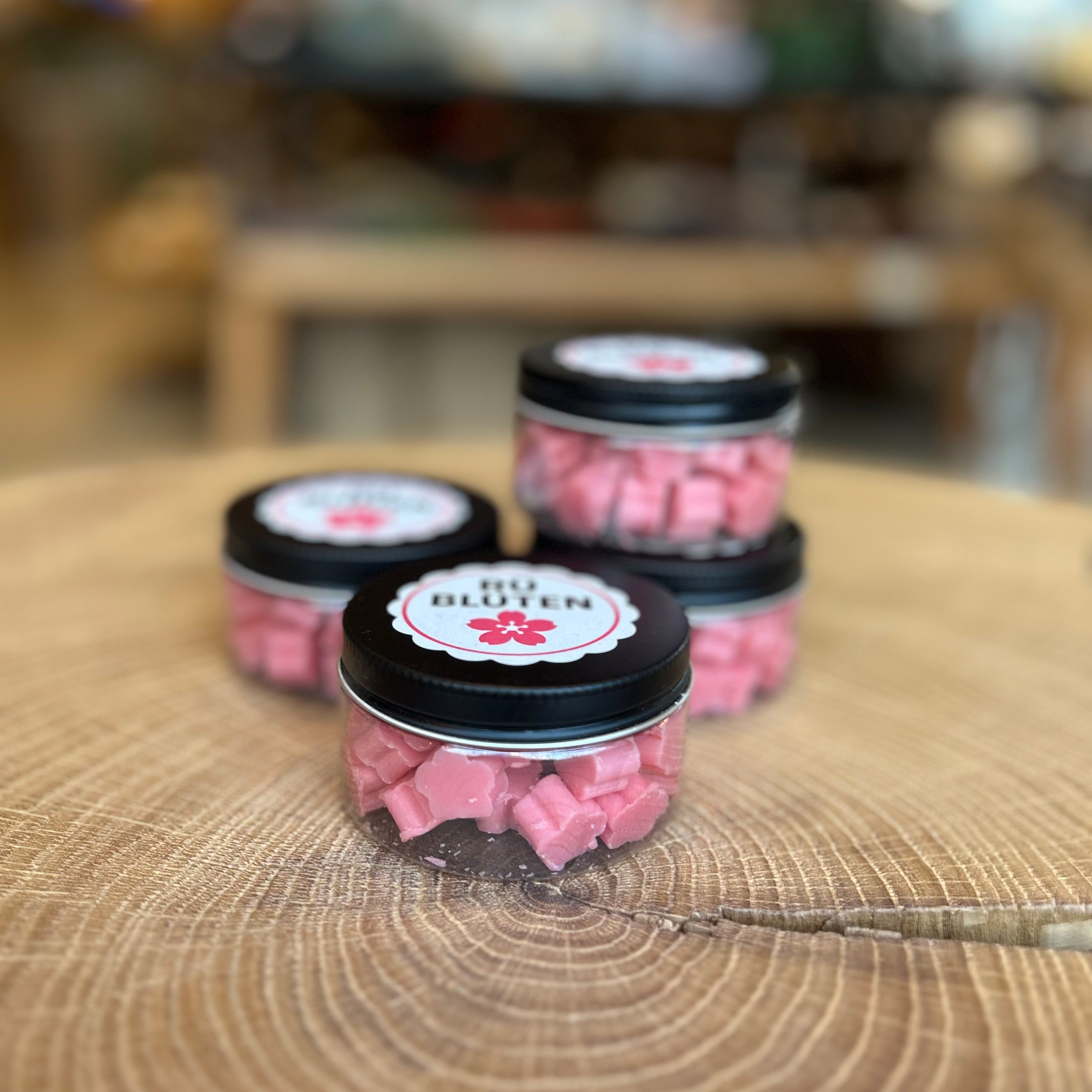 Kleine Dose mit rosa Seifenblüten „RÜ Blüten“ auf einem dekorativen Holztisch, im Hintergrund unscharfe Boutique-Atmosphäre. Handgemachte Mini-Seifenblüten als Geschenk, Dekoration oder Duftseife – ideal für Bad, Gäste-WC oder als Rüttenscheid-Souvenir.