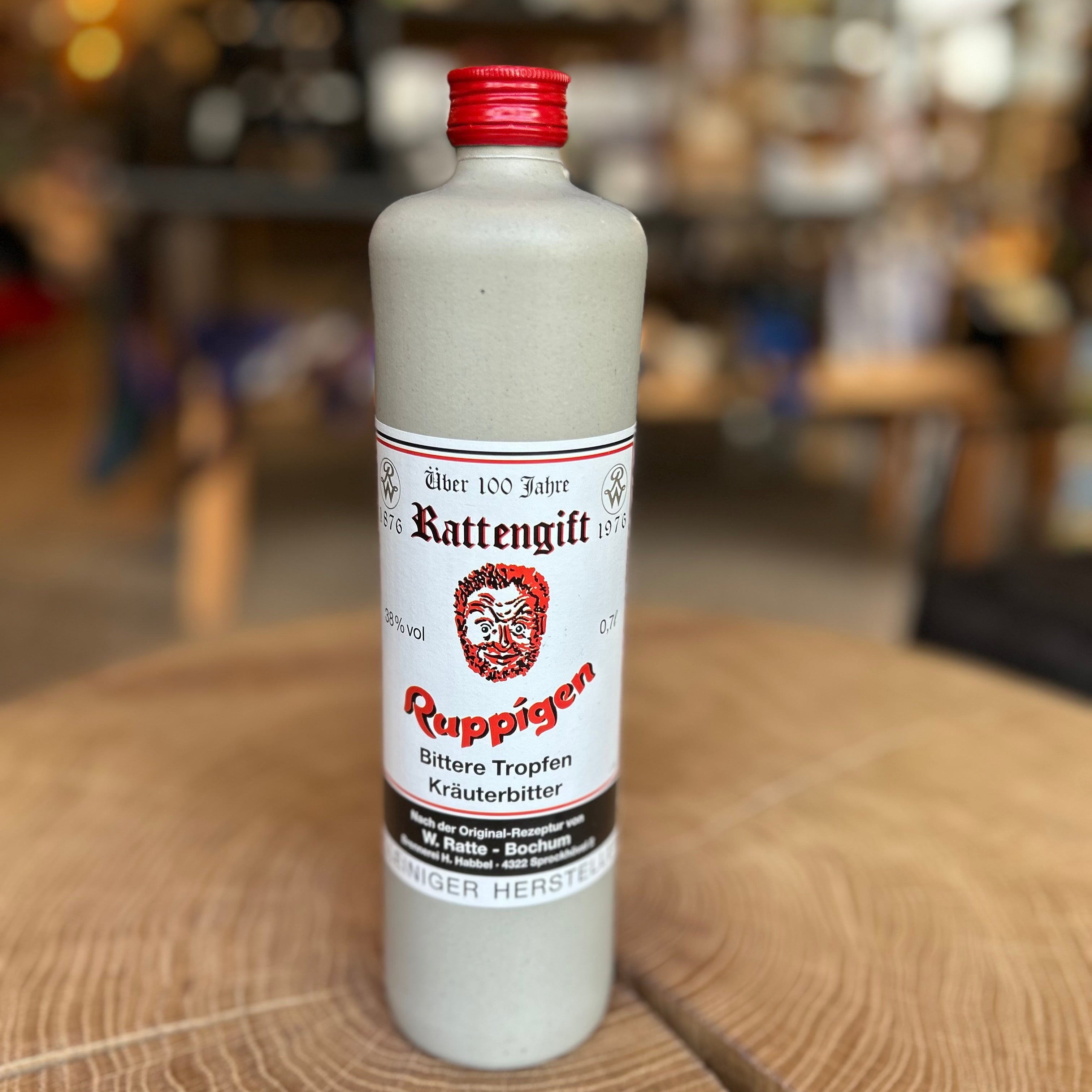 Steinzeugflasche mit rotem Schraubverschluss, Etikett ‚Ruppigen Bittere Tropfen Kräuterbitter – Rattengift‘, aufgenommen auf einem Holztisch mit unscharfem Hintergrund