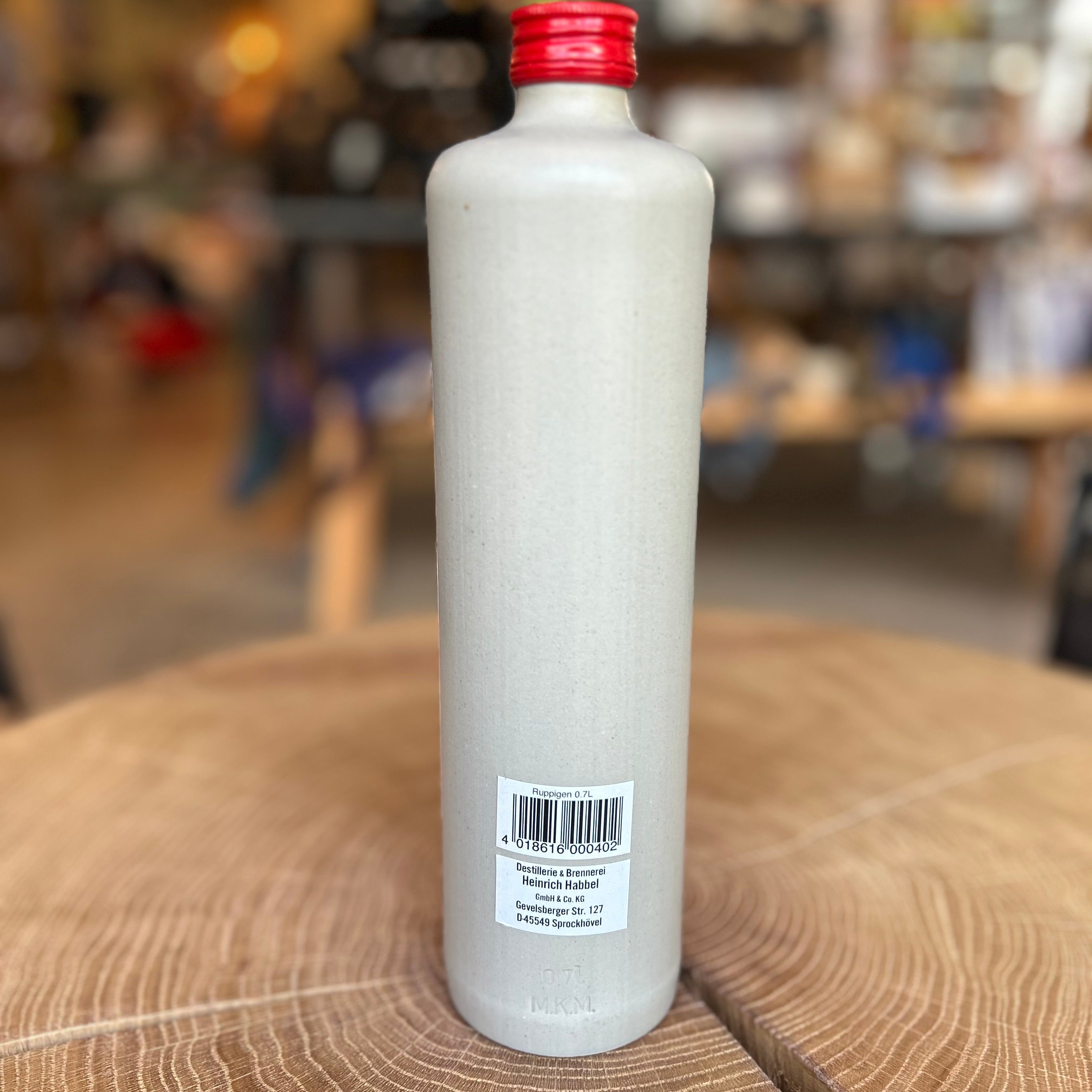 Steinzeugflasche von hinten mit rotem Schraubverschluss, Etikett ‚Ruppigen Bittere Tropfen Kräuterbitter – Rattengift‘, aufgenommen auf einem Holztisch mit unscharfem Hintergrund