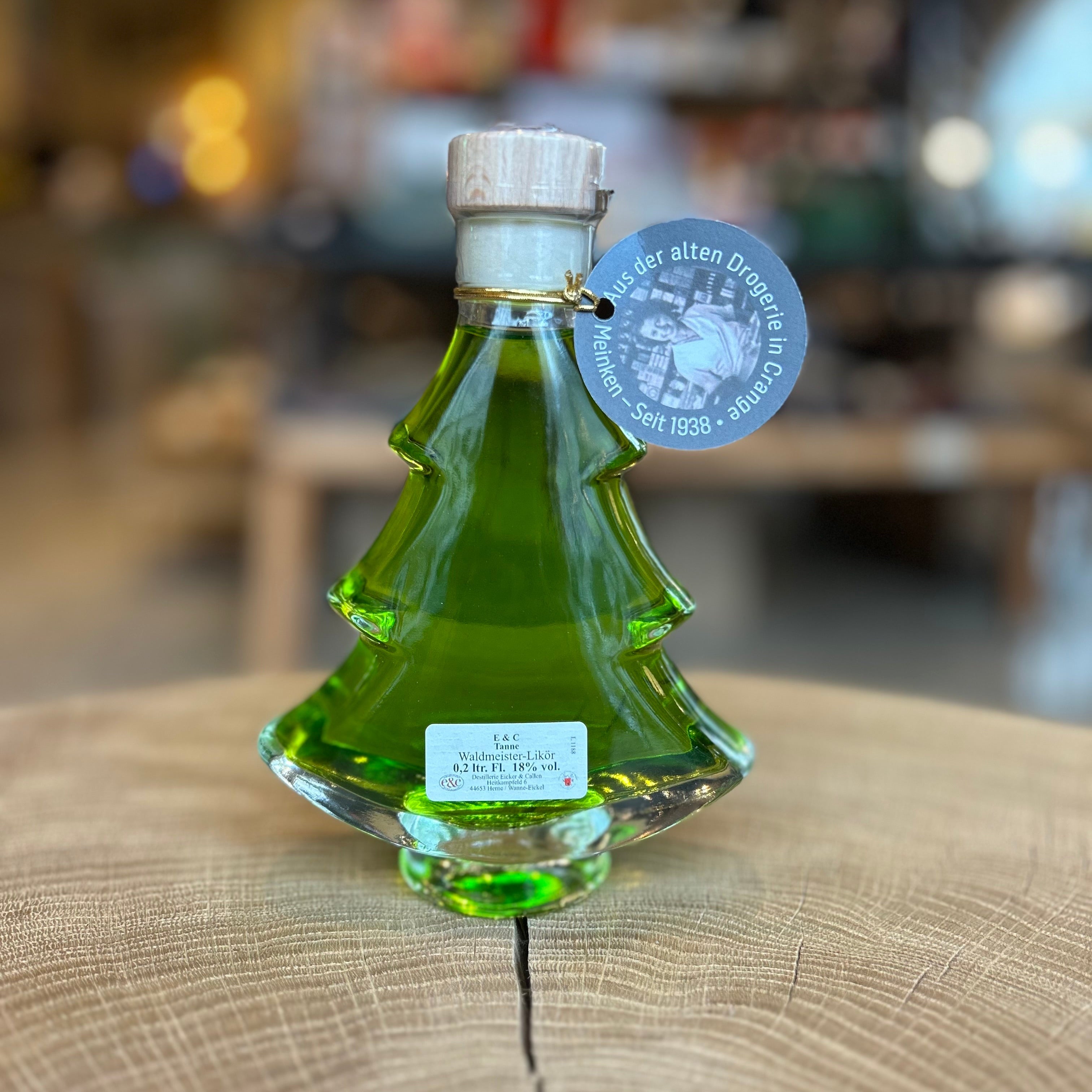 Grüne Likörflasche in Tannenbaumform, dekorativ auf einem Holztisch präsentiert. Weihnachtlicher Likör in besonderer Geschenkflasche aus Glas – ideal als Weihnachtsgeschenk, Wintergetränk oder festliches Mitbringsel. Hintergrund unscharf mit gemütlicher Ladenatmosphäre.
