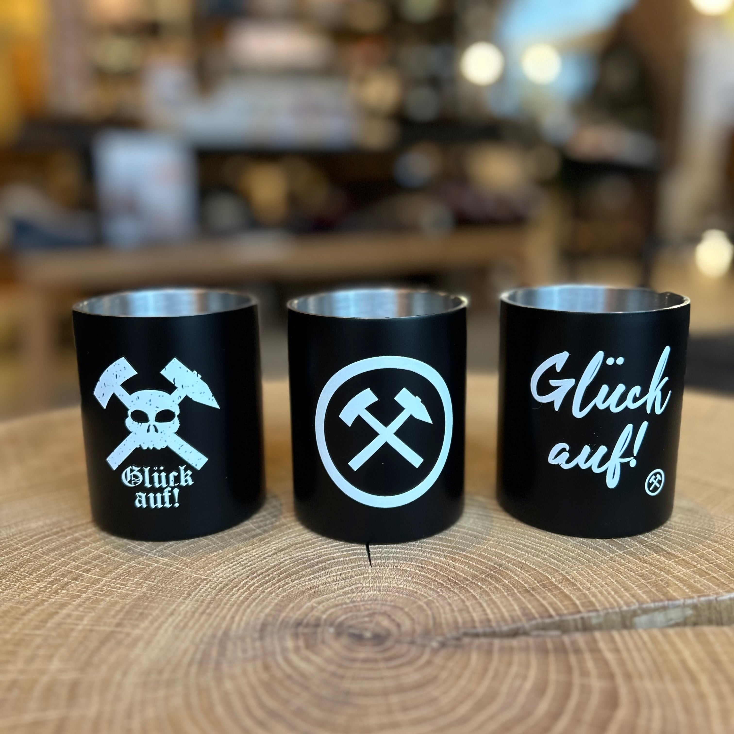 Schwarze Becher mit Ruhrpott-Motiven auf Holzoberfläche – drei Becher mit Bergbau-Symbolen, gekreuzten Hämmern und Schriftzug ‚Glück auf!‘, regionales Geschenk aus dem Ruhrgebiet