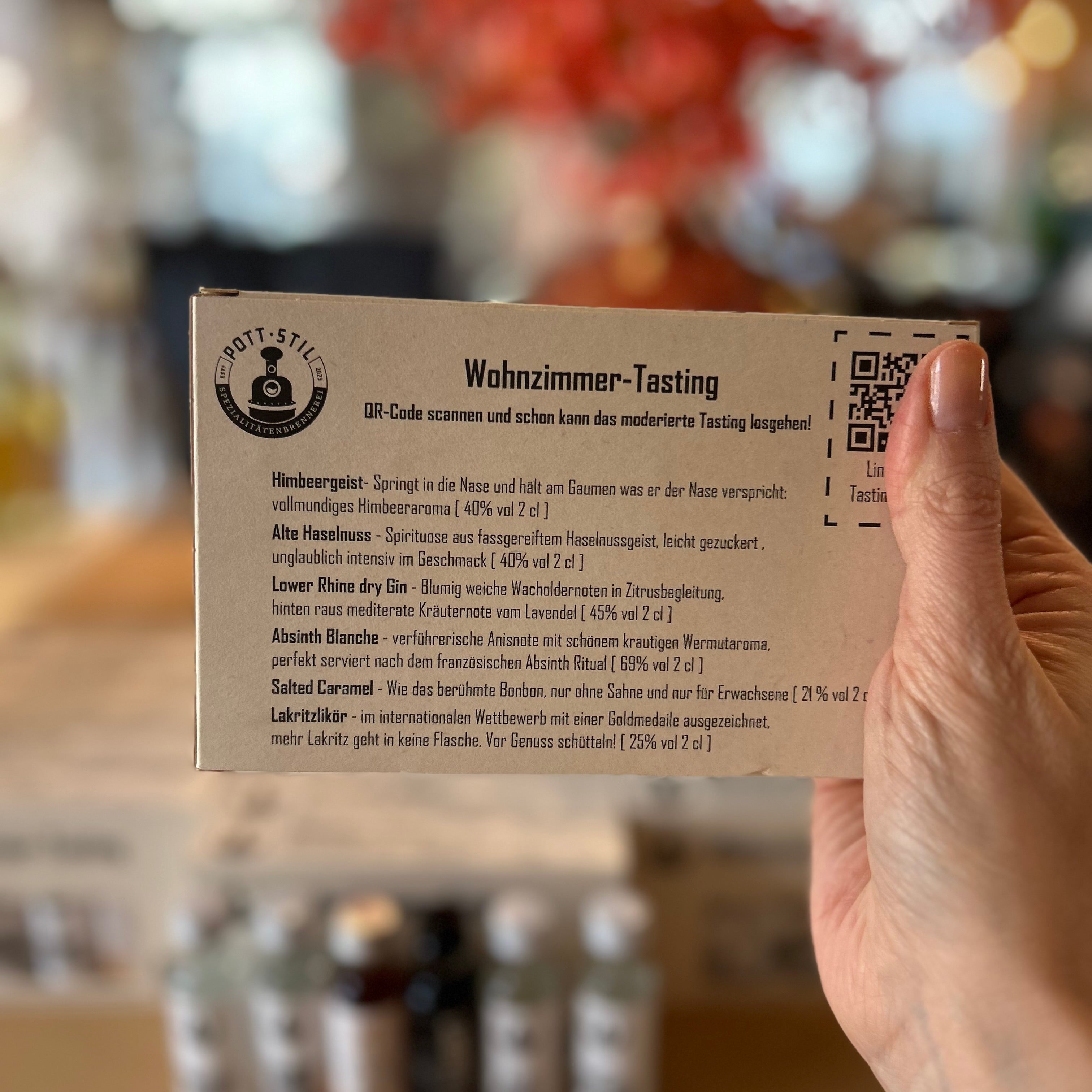 Wohnzimmer-Tasting Set mit verschiedenen Spirituosen-Miniaturen wie Himbeergeist, Gin, Lakritzlikör und Absinth auf einem Holzbrett vor Verpackungen des Wohnzimmer-Tasting Probierpakets – Spirituosen Verkostungsset für Zuhause.