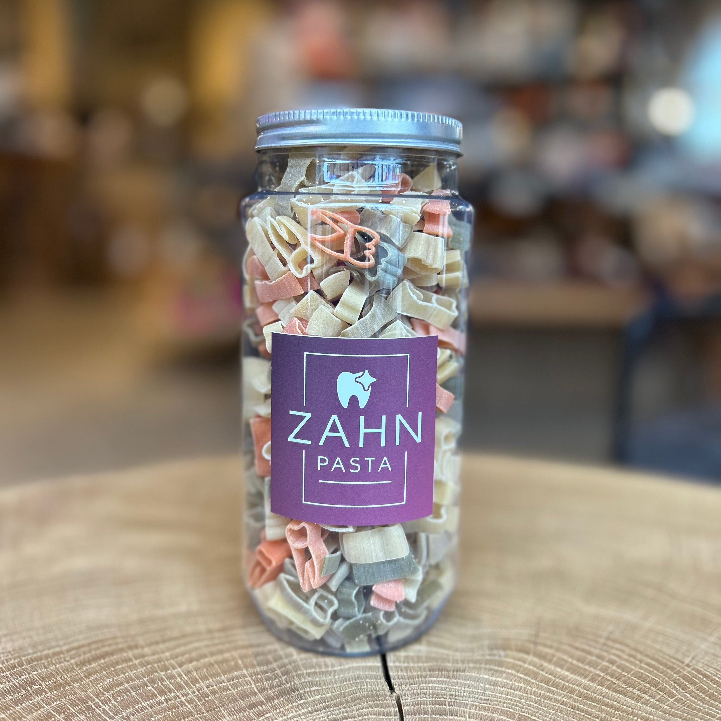 PET Glas mit farbiger „Zahn-Pasta“ in Zahnform auf einem Holzuntergrund. Das Produkt trägt ein violettes Etikett mit einem Zahn-Logo und der Aufschrift „ZAHN PASTA“. Ideal als Geschenk für Zahnärzte, Dentalpraxen oder Zahnpflege-Fans.